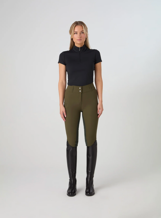 A woman stands against a light gray studio background wearing ps of sweden olive green fitted riding pants, a black short-sleeve mock-neck top, and tall black riding boots