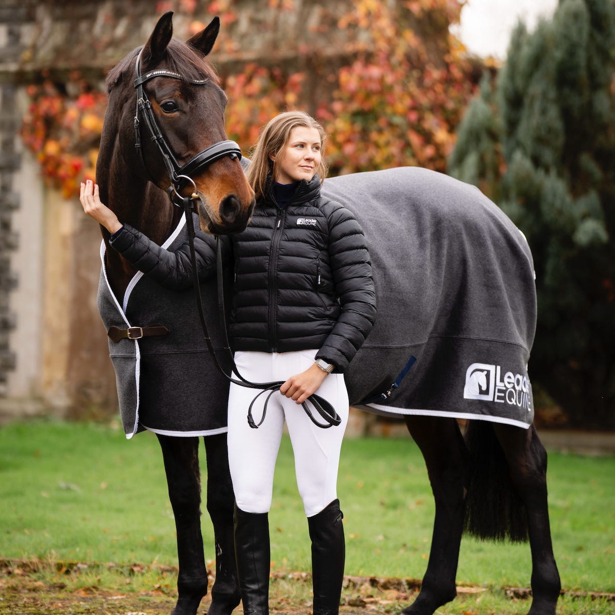Woman in Leader Equine equestrian attire standing next to a horse with a grey wool horse rug in an outdoor setting.