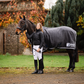 Woman in Leader Equine breeches and puffer jacket standing next to horse in grey wool rug with Leader Equine branding.
