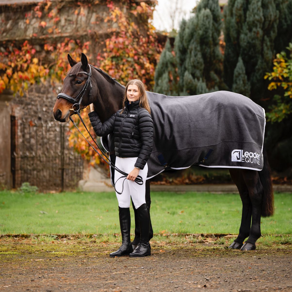 Woman in Leader Equine breeches and puffer jacket standing next to horse in grey wool rug with Leader Equine branding.