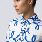 A close-up side profile of a woman wearing a white button-up shirt with a bold blue abstract pattern, set against a plain light gray studio background.