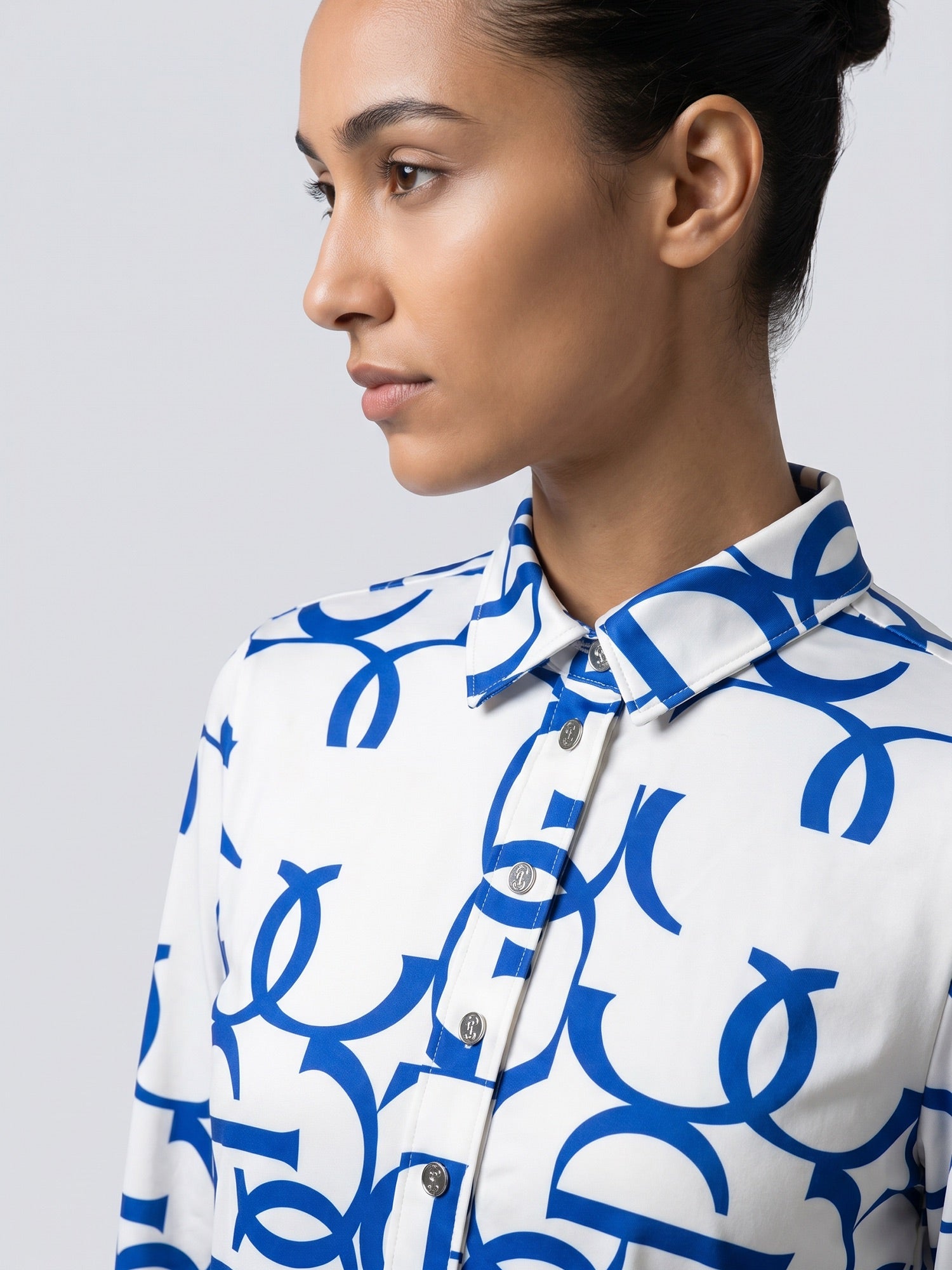A close-up side profile of a woman wearing a white button-up shirt with a bold blue abstract pattern, set against a plain light gray studio background.