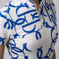 A close-up of a model wearing a short-sleeve athletic top with a white base and a bold blue abstract pattern, featuring a white half-zip collar.